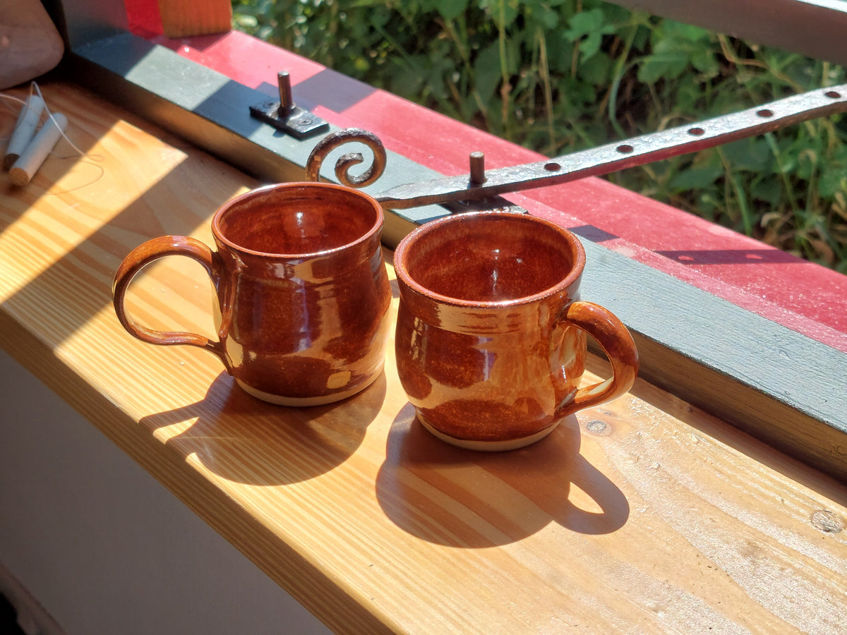 Two handmade terracotta ceramic mugs with a glossy brown-orange glaze, resting on a wooden windowsill in bright natural sunlight.