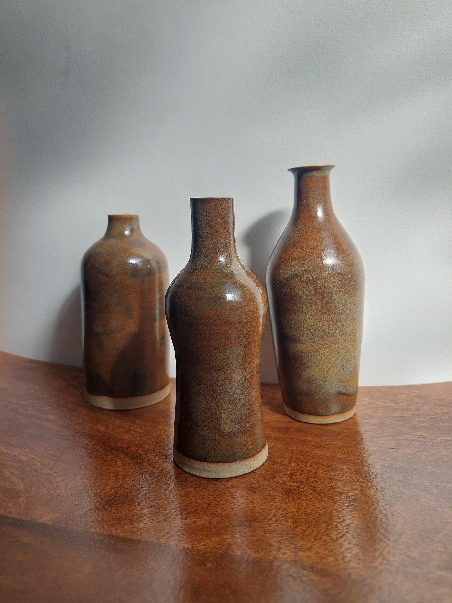 Three small brown ceramic bottles with narrow necks and light beige rims, arranged diagonally on a wooden surface against a white background.
