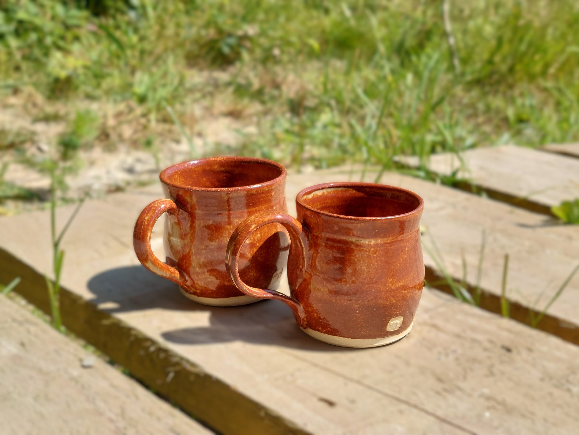 Two handmade ceramic mugs with a glossy terracotta-brown glaze and cream-colored bottoms, resting on a wooden deck with a blurred grassy background.
