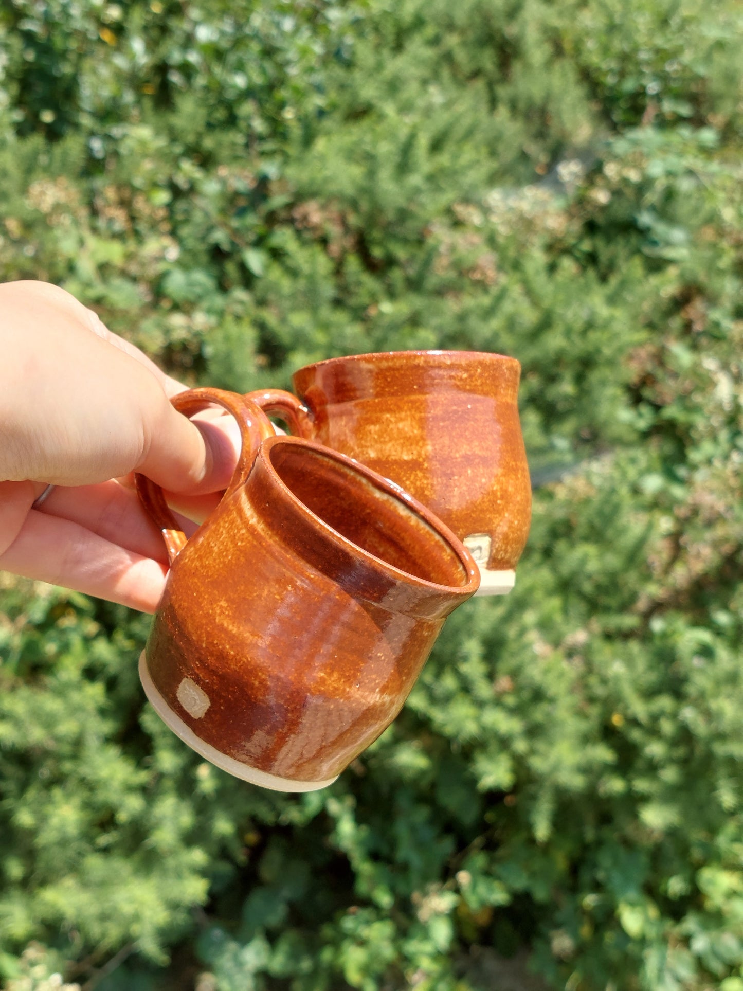 Two handmade ceramic mugs with a glossy terracotta-brown glaze are held by a hand against a blurred green outdoor background.