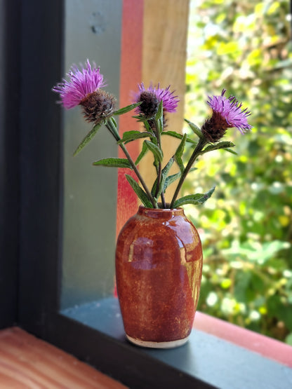 A small glazed ceramic vase with a warm reddish-brown finish holding three purple thistle flowers, placed on a windowsill with a blurred outdoor garden background.