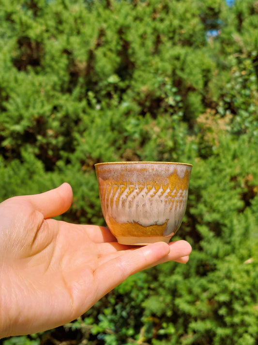 A handmade ceramic tea cup with a brown, gold, and white glaze, held in a person's hand against a blurred green outdoor background.