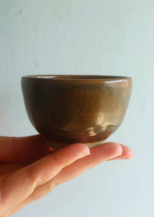 A small handmade ceramic bowl with a glossy dark brown and bronze glaze, held in a person's hand against a plain light gray wall.