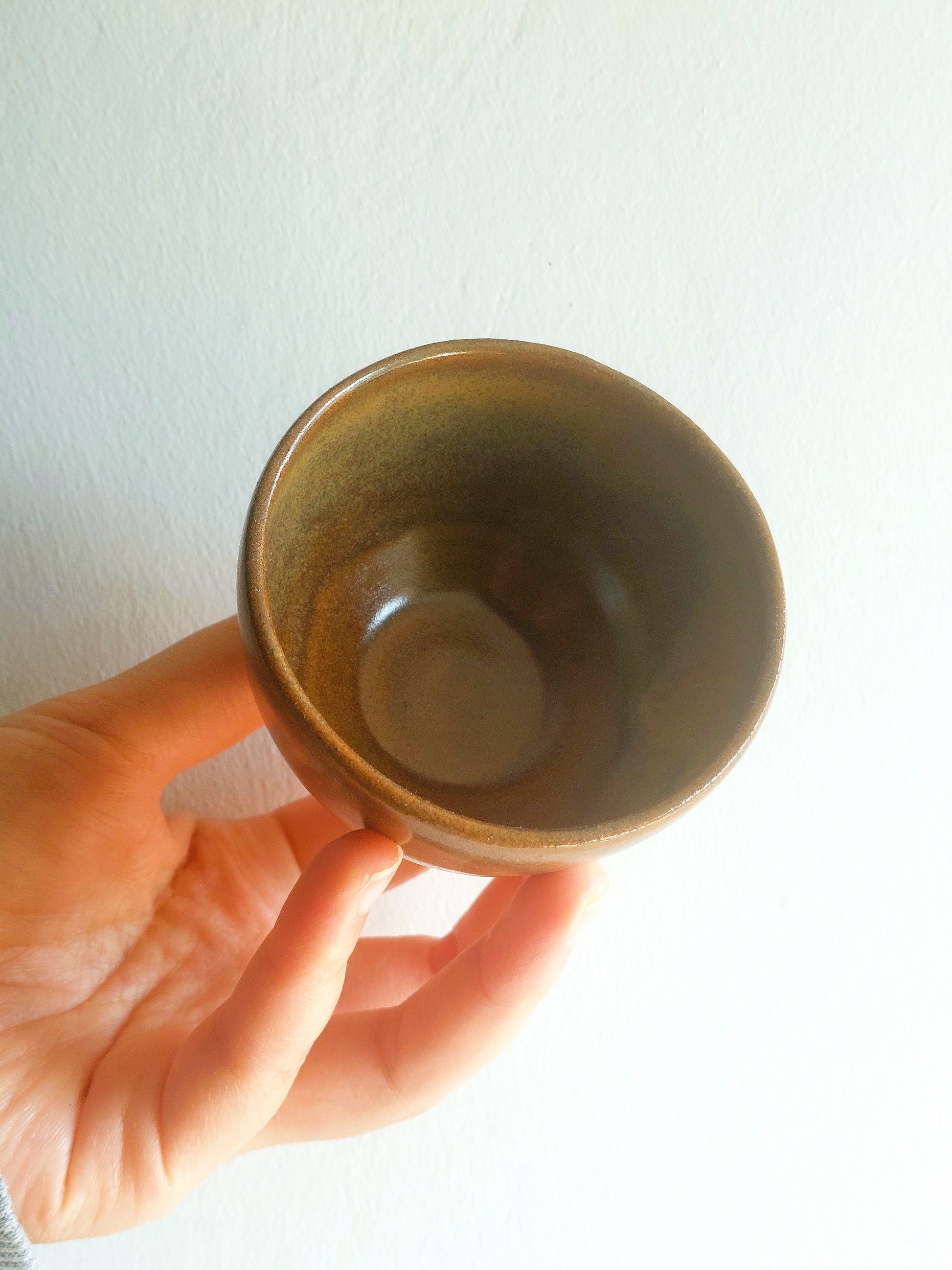 A hand holding a small handmade ceramic cup with a brown and olive-green glaze against a plain white wall.