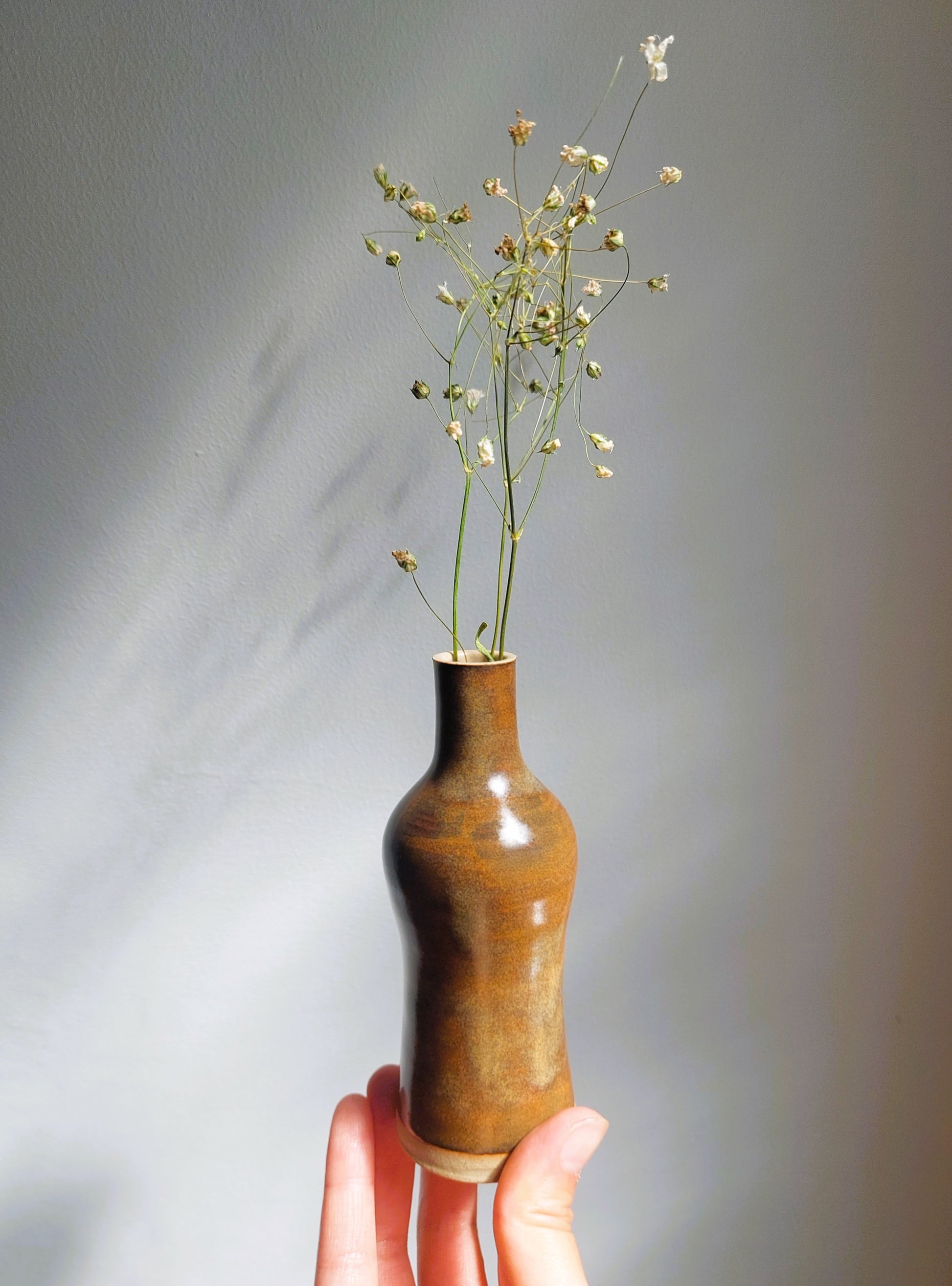 A small hand-held ceramic vase with a glossy brown finish, holding delicate white baby's breath flowers, held up by a hand against a neutral gray background.
