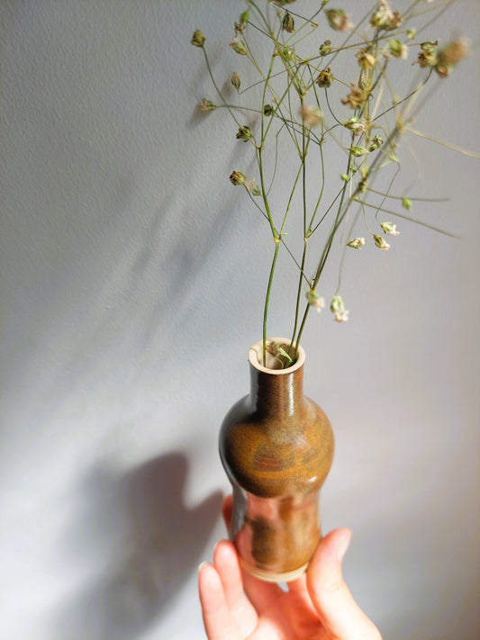 A hand holds a small brown ceramic bud vase filled with dried baby's breath flowers against a plain gray wall.