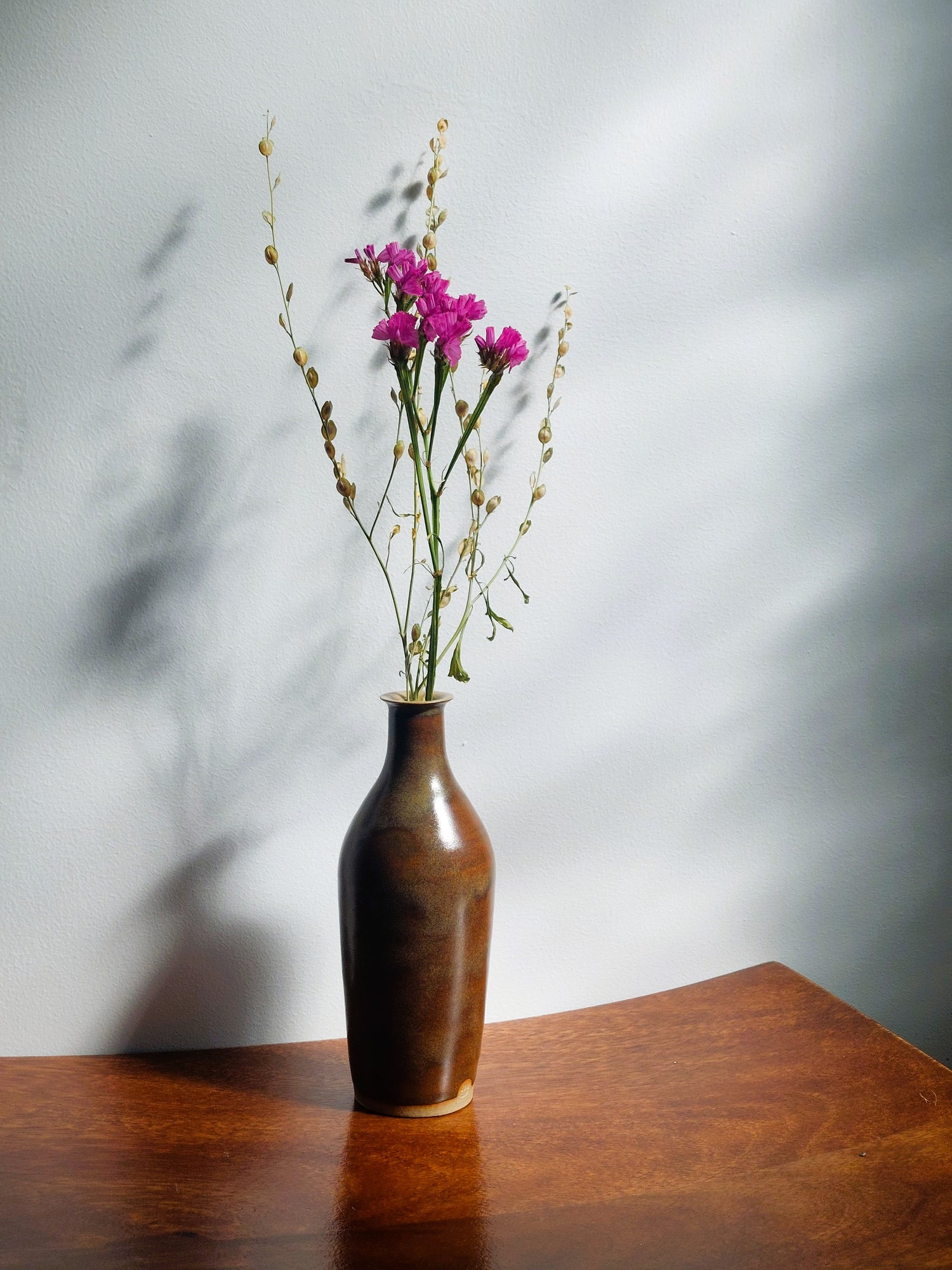 A brown ceramic vase with a glossy finish, holding dried pink flowers and green stems, set on a wooden table against a gray wall with soft shadows.