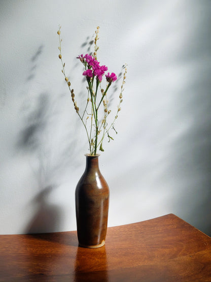 A brown ceramic vase with a glossy finish, holding dried pink flowers and green stems, set on a wooden table against a gray wall with soft shadows.