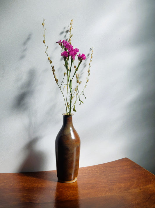 A brown ceramic vase with a glossy finish, holding dried pink flowers and green stems, set on a wooden table against a gray wall with soft shadows.