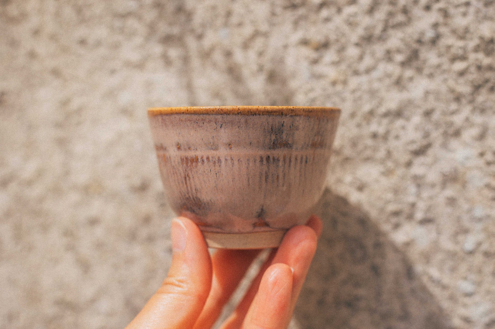 Hand holding a small ceramic cup against a textured stone wall.