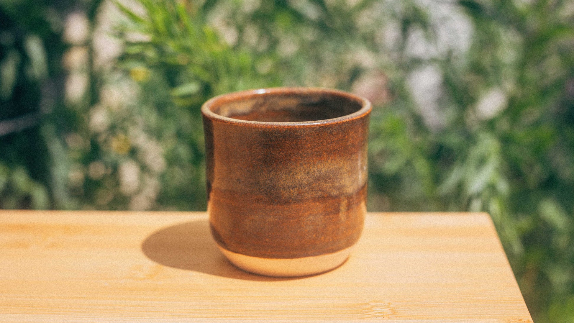 Brown ceramic cup on a wooden surface with a blurred green outdoor background