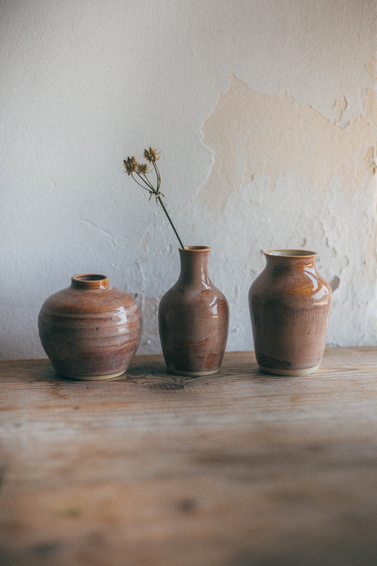 Three small brown ceramic vases with a glossy earthy glaze are lined up on a rustic wooden table against a textured off-white wall. The center vase holds a single dried flower stem.