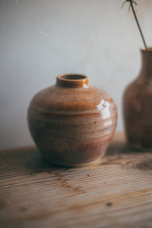 A small round ceramic vase with a glossy brown and amber glaze sits on a rustic wooden table against a soft light gray wall. A second blurred vase is visible in the background on the right.