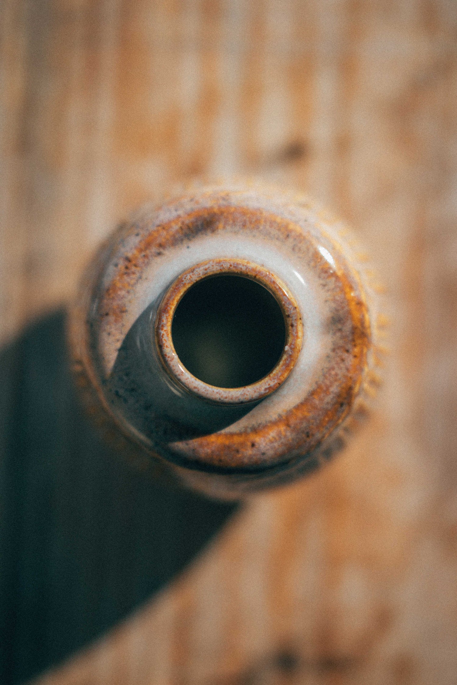 A close-up, top-down view of a ceramic bottle or vase opening with a textured, glazed finish in earthy brown, beige, and white tones, set against a rustic wooden background.