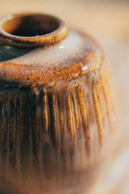 Close-up of a small ceramic vase with a narrow opening, featuring a textured vertical ribbed pattern and a glossy brown and amber glaze.